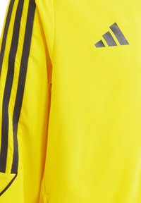 Gelbe sportliche Jacke mit schwarzen Seitenstreifen und Adidas-Logo. Hergestellt aus glattem, leichtem Material mit einer strukturierten Passform und ohne sichtbare Muster.
