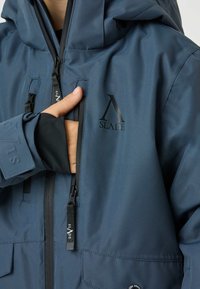 Giacca impermeabile blu con cappuccio, dotata di chiusure con cerniera, un logo e polsini con foro per il pollice. Tessuto liscio, tasche pratiche.