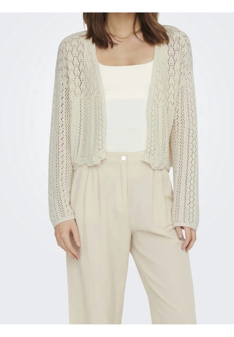Jasnobeżowy krótki kardigan z ażurowej dzianiny, z długimi rękawami i otwartym przodem. W połączeniu z białym topem i beżowymi spodniami.