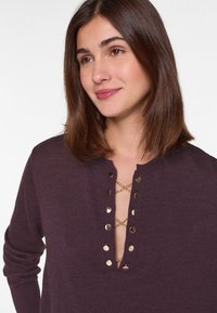 Bordeauxfarbener Pullover mit tiefem Ausschnitt, verziert mit Akzenten in Goldtönen und einer Kettendetaillierung. Weicher Stoff und lange Ärmel.