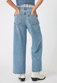 Jeans de pierna ancha en un tono azul claro con una cintura alta, dos bolsillos traseros y detalles de costura sutiles. Combinados con botas blancas hasta el tobillo.