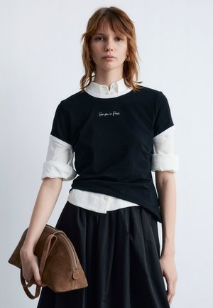 Black t-shirt layered over a white collared shirt, featuring embroidered text. Paired with a dark skirt and holding a brown suede handbag.