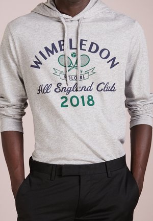 Personne portant un sweat à capuche gris clair avec le texte « Wimbledon All England Club 2018 » et un graphisme de raquettes de tennis croisées sur le devant.