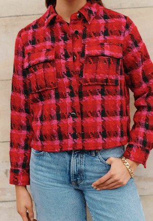Red and pink plaid jacket with black accents, featuring two chest pockets, a pointed collar, and buttons down the front, paired with blue jeans.