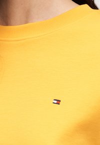 Κίτρινο βαμβακερό φούτερ με στρογγυλή λαιμόκοψη και μικρό, κεντημένο λογότυπο με κόκκινες, λευκές και μπλε λεπτομέρειες στο στήθος.