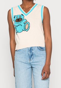 Ärmellös krämfärgad stickad tröja med en blå björngrafik, med turkos kant längs v-ringningen och ärmhålen.