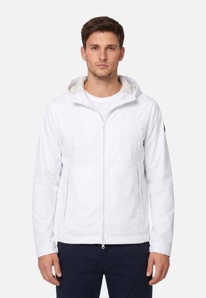 Giovane uomo che indossa una giacca con cappuccio bianca con cerniera e pantaloni scuri, in piedi davanti a uno sfondo chiaro uniforme.