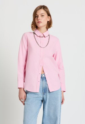 Frau trägt ein hellrosa Hemd mit Knöpfen, eine silberne Kettenkette und hellblaue, hoch taillierte Jeans vor einem schlichten Hintergrund.