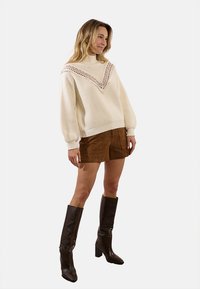 Maglione crema con un accento in pizzo a forma di V, abbinato a una mini gonna in suede marrone e stivali alti neri con tacco. Tessuto morbido, vestibilità over.