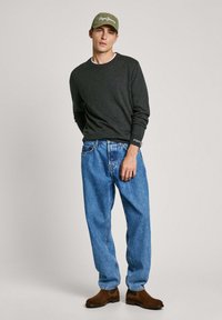 Tmavě šedý svetr s dlouhým rukávem, světlý šedý límec, modré volné džíny, hnědé kotníkové boty, zelená čepice s bílým logem, jednobarevné pozadí.