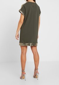 Zielona oliwkowa sukienka z krótkimi rękawami, złotym wykończeniem z cekinów na rękawach i u dołu, z kluczykowym wycięciem z tyłu. Połączona z srebrnymi sandałami na wysokim obcasie.