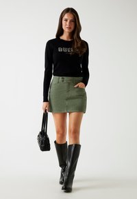 Zwarte langslavende T-shirt met "QUEST" graphic, groene denim rok en zwarte kniehoge laarzen. Houdt een kleine zwarte handtas vast.
