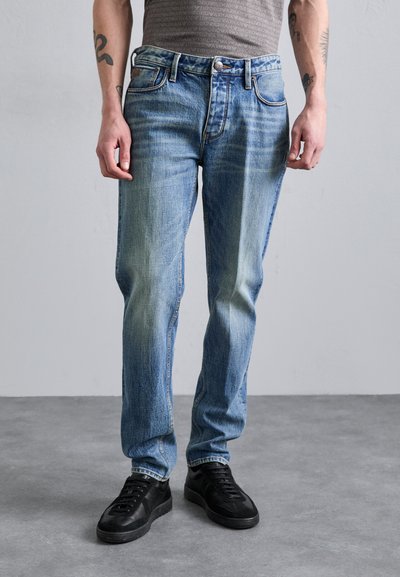 Jean en denim bleu clair avec une coupe régulière, présentant une légère décoloration, un design à cinq poches et des accessoires en métal. Associés à des baskets noires.