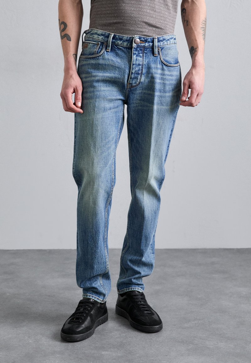 Light blue denim jeans with a regular fit, featuring subtle fading, five-pocket design, and metal hardware. Paired with black sneakers.