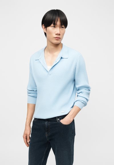 Jeune homme aux cheveux noirs portant un t-shirt à manches longues côtelé bleu clair et un jean foncé, debout avec une main dans la poche.