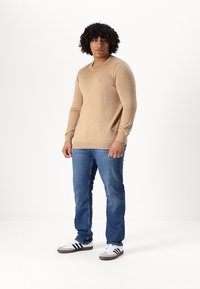 Camisa beige de manga larga con cuello redondo, combinada con jeans azules y zapatillas blancas con detalles en negro. Diseño sencillo y ajustado.