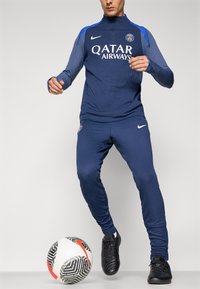 Navy blue athletic tracksuit with ribbed texture and a half-zip collar. Features white "QATAR AIRWAYS" logo and black sneakers, next to a patterned soccer ball.