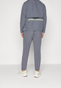 Grå atletisk träningsoverall med huva, logotypdetaljer på ryggen och smala byxor. Texturerad tyg med elastiska mudd och midja.