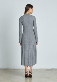Abito midi grigio a coste con maniche lunghe, texture plissè, collo alto e una leggera lucentezza; abbinato a sandali neri con tacco.
