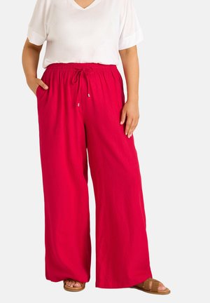 Person trägt leuchtend rote, weit geschnittene Hosen mit elastischem Kordelzugbund, weißes Kurzarmhemd und braune Sandalen mit offenen Zehen.