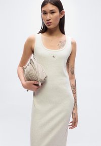 Αμάνικο φόρεμα ανοιχτού γκρι με εφαρμοστή γραμμή, διακοσμημένο με ένα λογότυπο. Κρατώντας μια τσάντα ώμου σε χρώμα ταουπέ με πτυχωτή υφή.