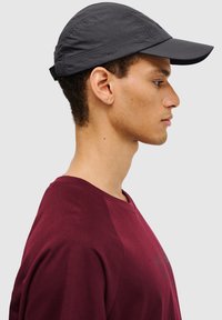 Schwarze leichte Kappe mit starrem Schirm, aus strukturiertem Stoff und mit dem Label "OFF PITCH" an der Seite. Getragen von einer Person in einem burgunderfarbenen Hemd.