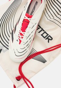 White soccer shoe with red accents and black details, featuring a textured upper and sleek design. Accompanied by a white drawstring bag with black lines.