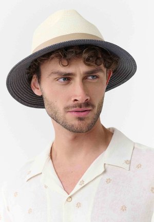 Man with curly hair wearing a beige and black wide-brim hat and a light cream shirt with subtle floral patterns, looking to the side.