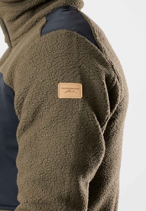 Fleece-takki oliivinvihreänä, mustilla nylonsomisteilla harteilla. Varustettu vaaleanruskealla nahkatunnuksella, jossa on "SWEDEMOUNT"-logo. Karhea pinta.