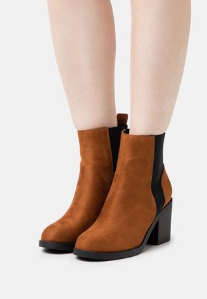 Bottes chelsea en suède marron avec un large talon noir et des panneaux latéraux élastiques. Texture lisse avec un bout arrondi et une languette à l'arrière.