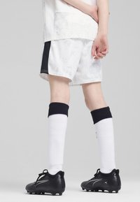 Pantalones cortos deportivos blancos con sutiles patrones grises y acentos laterales negros, combinados con zapatos de fútbol negros y calcetines blancos hasta la rodilla.