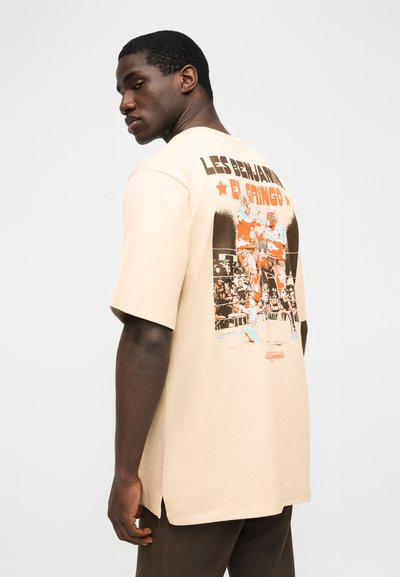 Άνδρας που φοράει μπεζ μπλούζα με σχέδιο δύο παλαιστών και το κείμενο "Les Benjamins El Gringo" στην πλάτη, στέκεται μπροστά σε απλό φόντο.