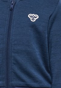 Granatowy sweter zapinany na zamek z małym haftowanym logo pszczoły po lewej stronie klatki piersiowej.