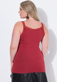 Žena s dlouhými blond vlasy, oblečená v červeném topu bez rukávů s krajkovým lemem a černé sukni, stojící zády k bílé zdi.