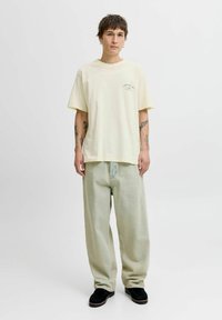 Gaiši dzeltena pārspīlēta T-kreklu ar mazu zaļu grafiku, apvienots ar gaiši zaļām platajām biksēm un melnajām kurpēm. Minimalistisks dizains.