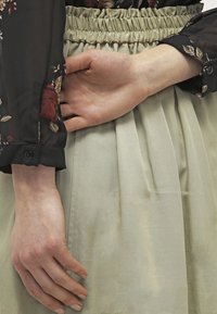Blusa nera con stampa floreale e maniche lunghe in trasparenza; gonna verde chiaro con pieghe e vita elastica. Mano appoggiata sulla vita.