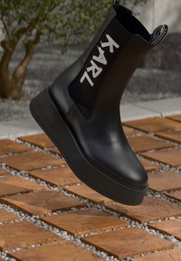 Botte plate en cuir noir avec panneau latéral élastique affichant un lettrage « KARL » en blanc, flottant au-dessus de carreaux carrés en bois avec des galets gris.