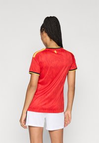 Γυναίκα με πλεγμένα μαλλιά, ντυμένη με κόκκινη αθλητική μπλούζα με κίτρινες και μαύρες λεπτομέρειες και λευκές βερμούδες, η οποία στέκεται με την πλάτη στραμμένη προς την κάμερα σε απλό φόντο.