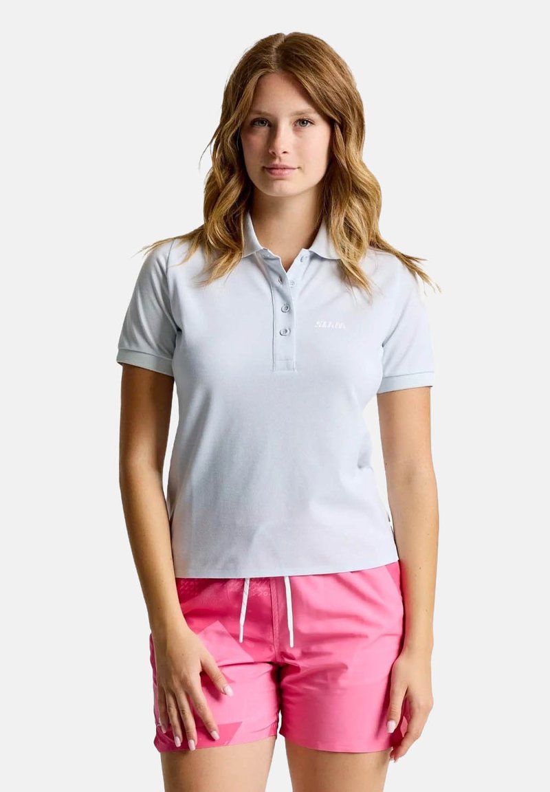Hellblaues Poloshirt mit einer Knopfleiste mit drei Knöpfen, kurzen Ärmeln und einer weichen Textur, kombiniert mit rosa Shorts mit geometrischen Mustern.