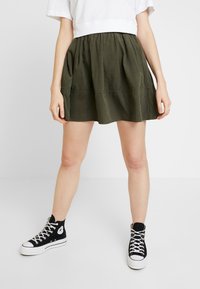 Jupe vert olive avec un tissu froncé, longueur mi-cuisse, présentant une coupe décontractée et des détails de couture subtils, assortie à des baskets montantes noires.