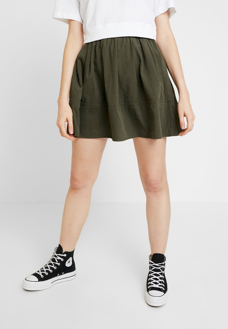 Jupe vert olive avec un tissu froncé, longueur mi-cuisse, présentant une coupe décontractée et des détails de couture subtils, assortie à des baskets montantes noires.