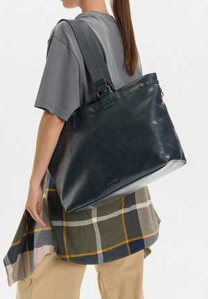 Žena nese velkou tmavou koženou tašku přes rameno s širokým popruhem, oblečená v šedé košili a kostkované sukni přes béžové kalhoty.