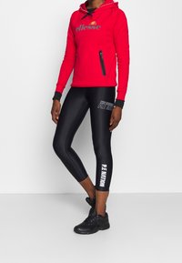 Personne portant un sweat à capuche rouge de la marque ellesse et des leggings noirs P.E NATION avec des baskets noires, debout devant un fond blanc.