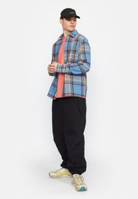 Blauwe geruite flanellen shirt over een koraalkleurig T-shirt, gecombineerd met zwarte baggy broek en beige sportschoenen met blauwe accenten. Zwarte pet inbegrepen.