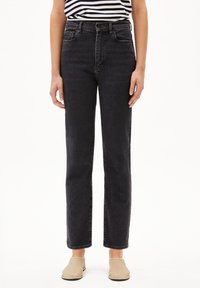 Jean noir taille haute en denim, avec cinq poches, une fermeture éclair et une coupe droite. Porté avec un haut rayé et des chaussures beiges.