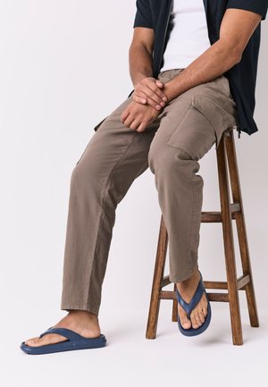 Homme portant un pantalon cargo beige et des tongs bleues, assis sur un tabouret en bois avec les mains jointes sur les genoux, sur fond blanc.