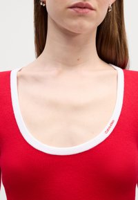 Rød ribbet topp med hvit rund hals, med en Calvin Klein-logo på kanten. Glatt tekstur med tettsittende design.