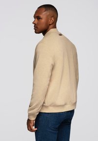 Beżowy sweatshirt o teksturowanym wykończeniu i okrągłym dekolcie, z logo na plecach. Połączony z ciemnoniebieskimi dżinsami.