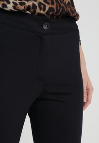 Black fitted pants made of a smooth, stretchy fabric, featuring a front zipper pocket and a matching button closure at the waist.
