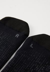 Schwarze Socken aus strukturiertem Stoff, mit "R" und "L" für den rechten und linken Fuß gekennzeichnet, mit einem einfarbigen oberen Teil und einem gemusterten Körper.